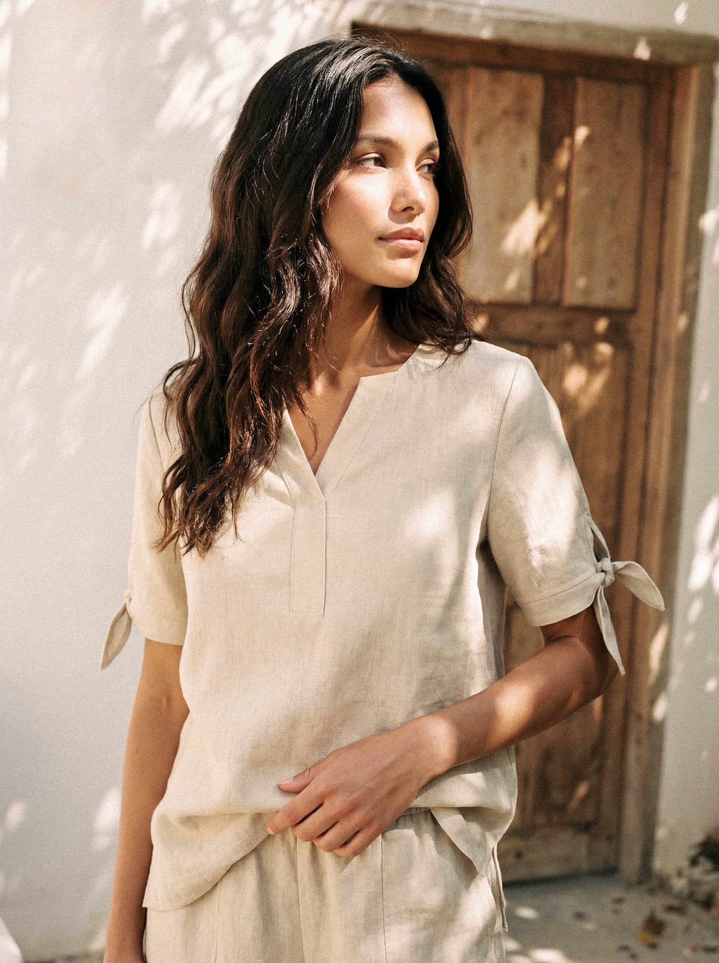 Woman wearing a light-colored outfit standing outdoors with a wooden door in the background