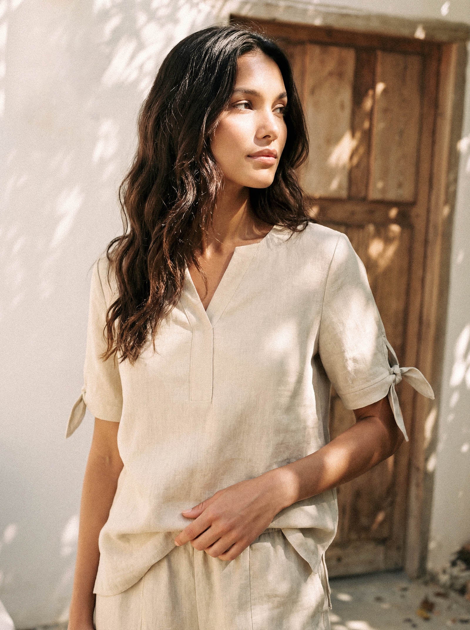 Woman wearing a light-colored outfit standing outdoors with a wooden door in the background