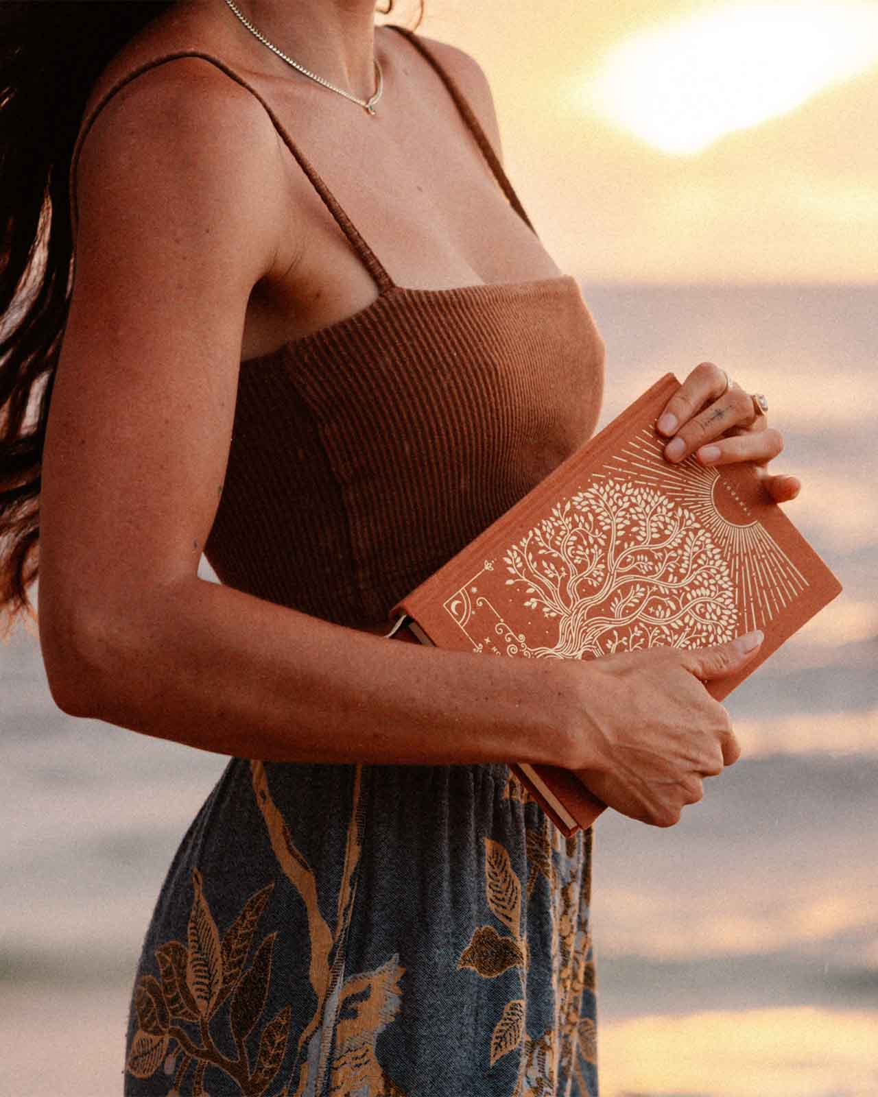 Woman holding a book with a tree design against a sunset beach backdrop