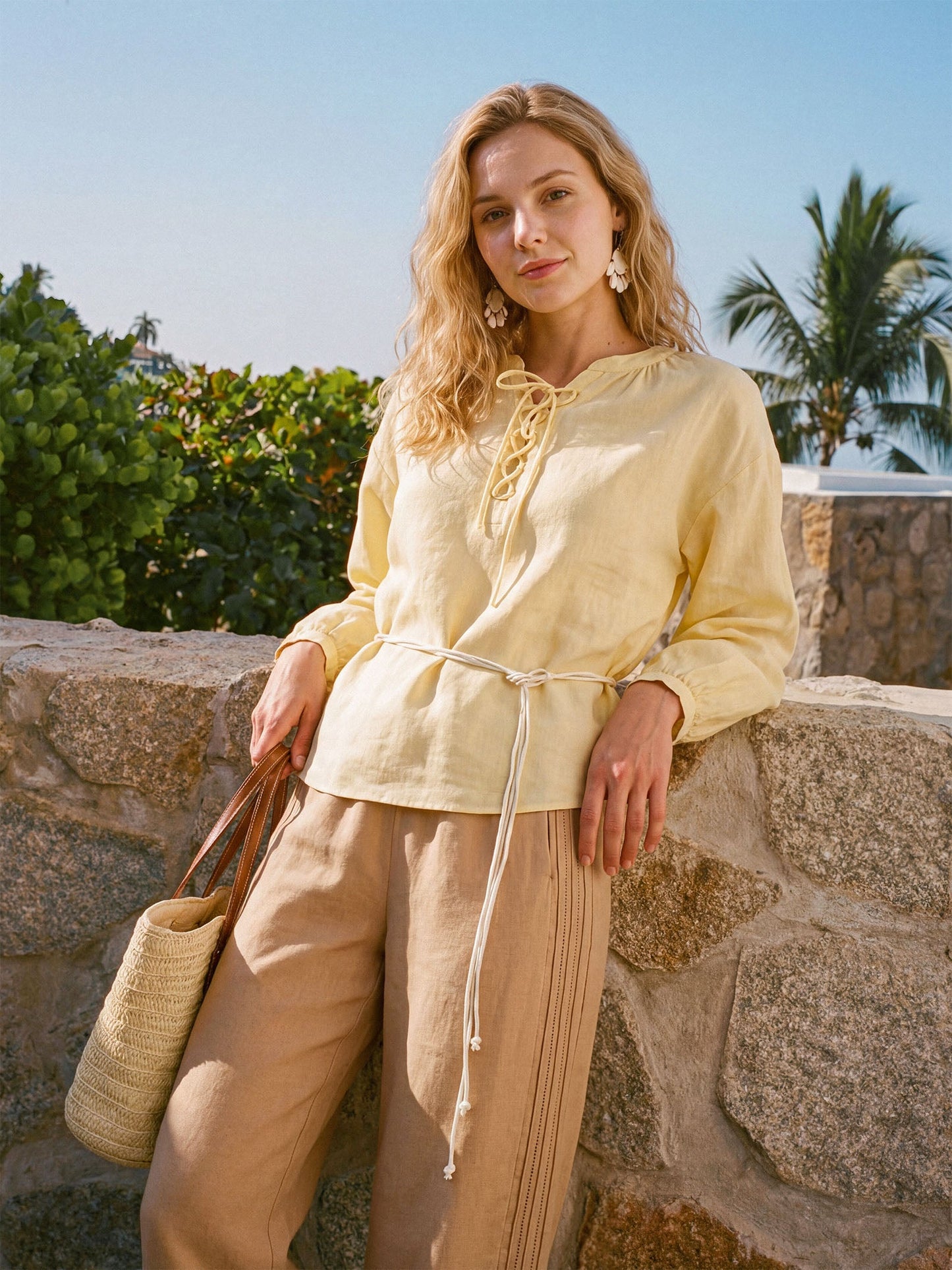 Woman in a yellow top and beige pants standing against a stone wall with palm trees in the background
