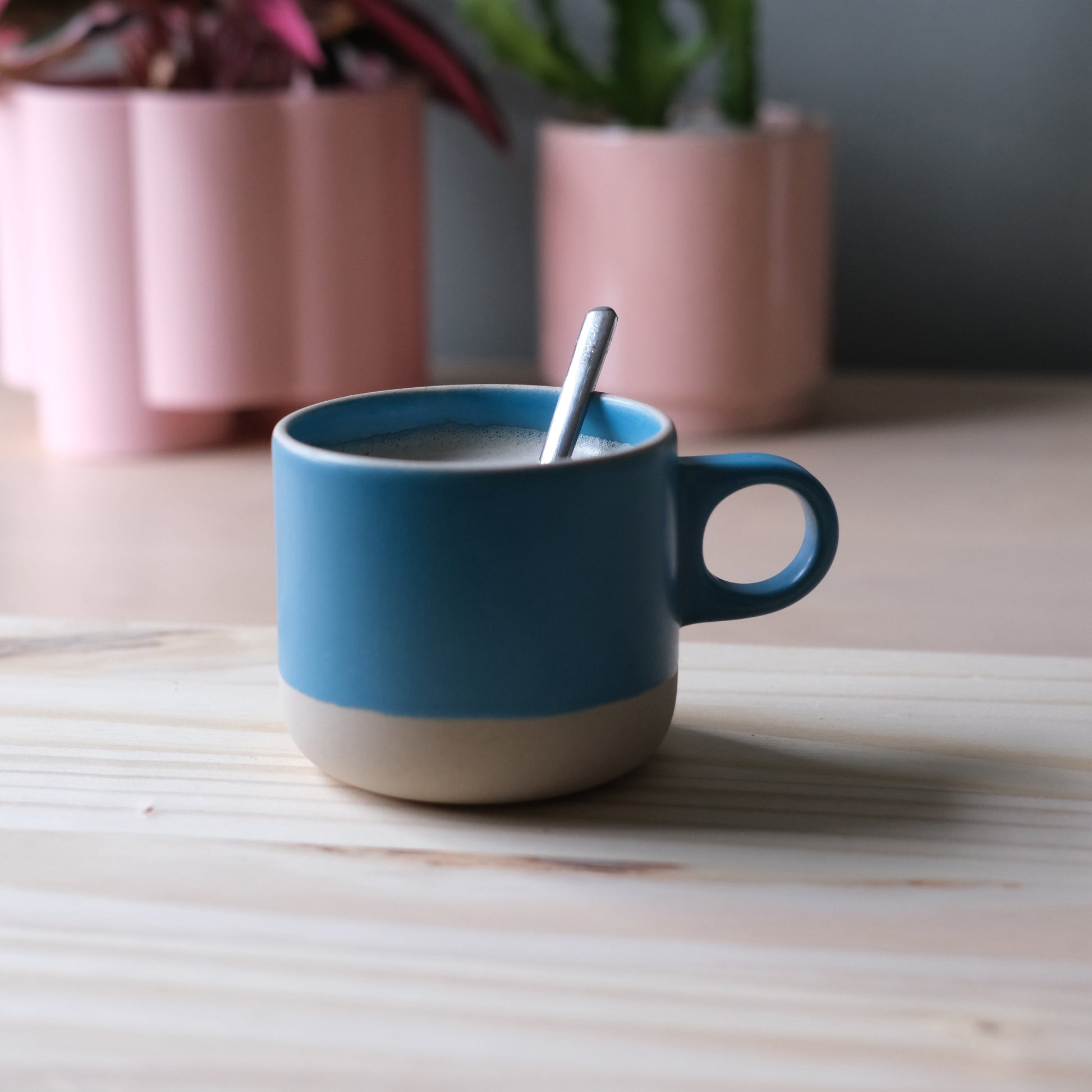 Blue mug with beige base on a wooden surface, surrounded by pink plant pots.