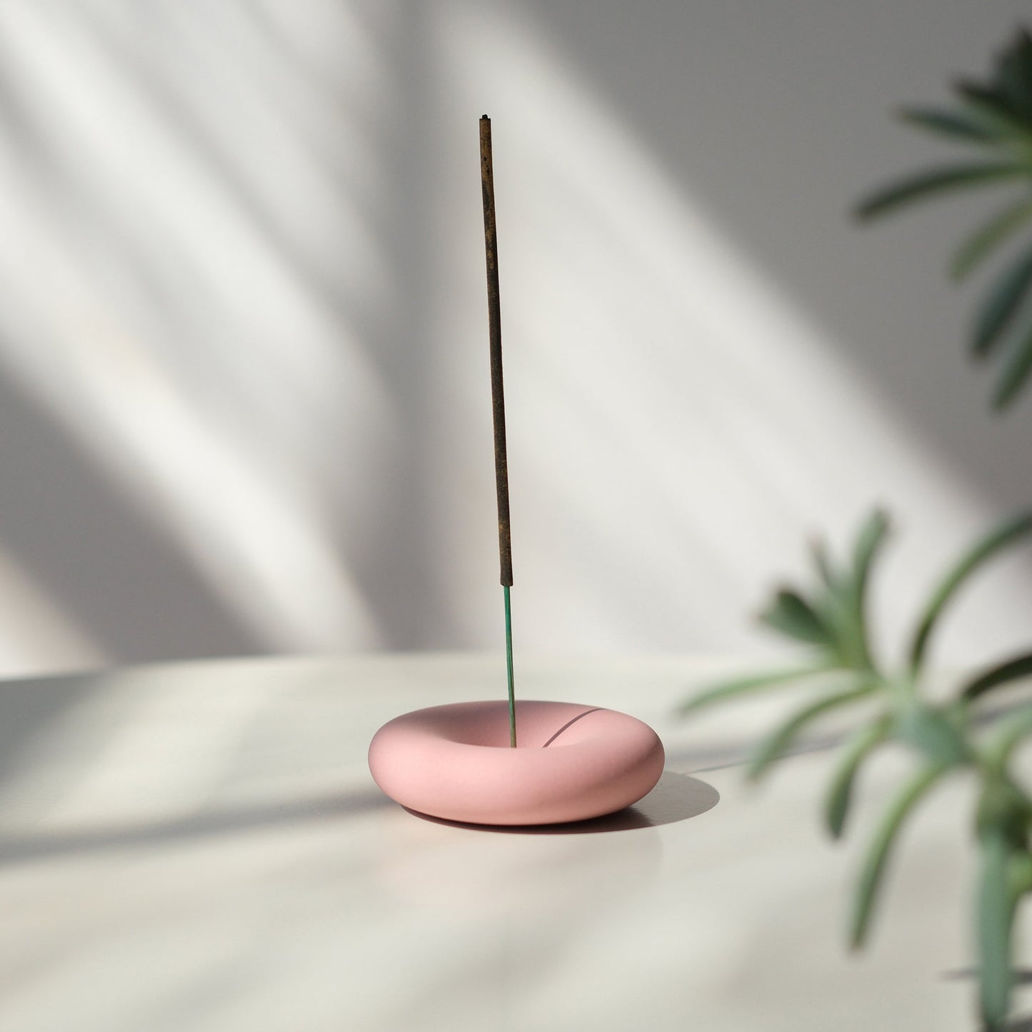 Pink incense burner with a single stick on a light surface with a plant in the corner