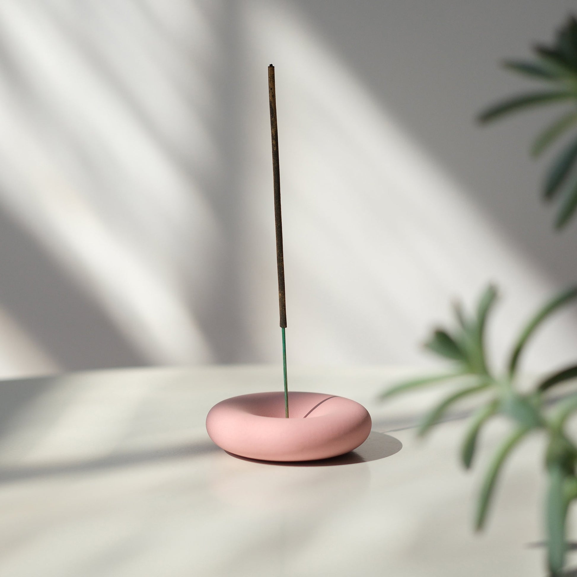 Pink incense burner with a single stick on a light surface with a plant in the corner