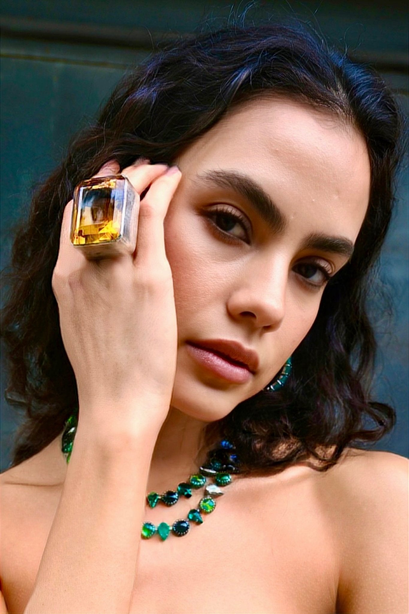 Woman wearing a large diamond ring and green necklace against a neutral background