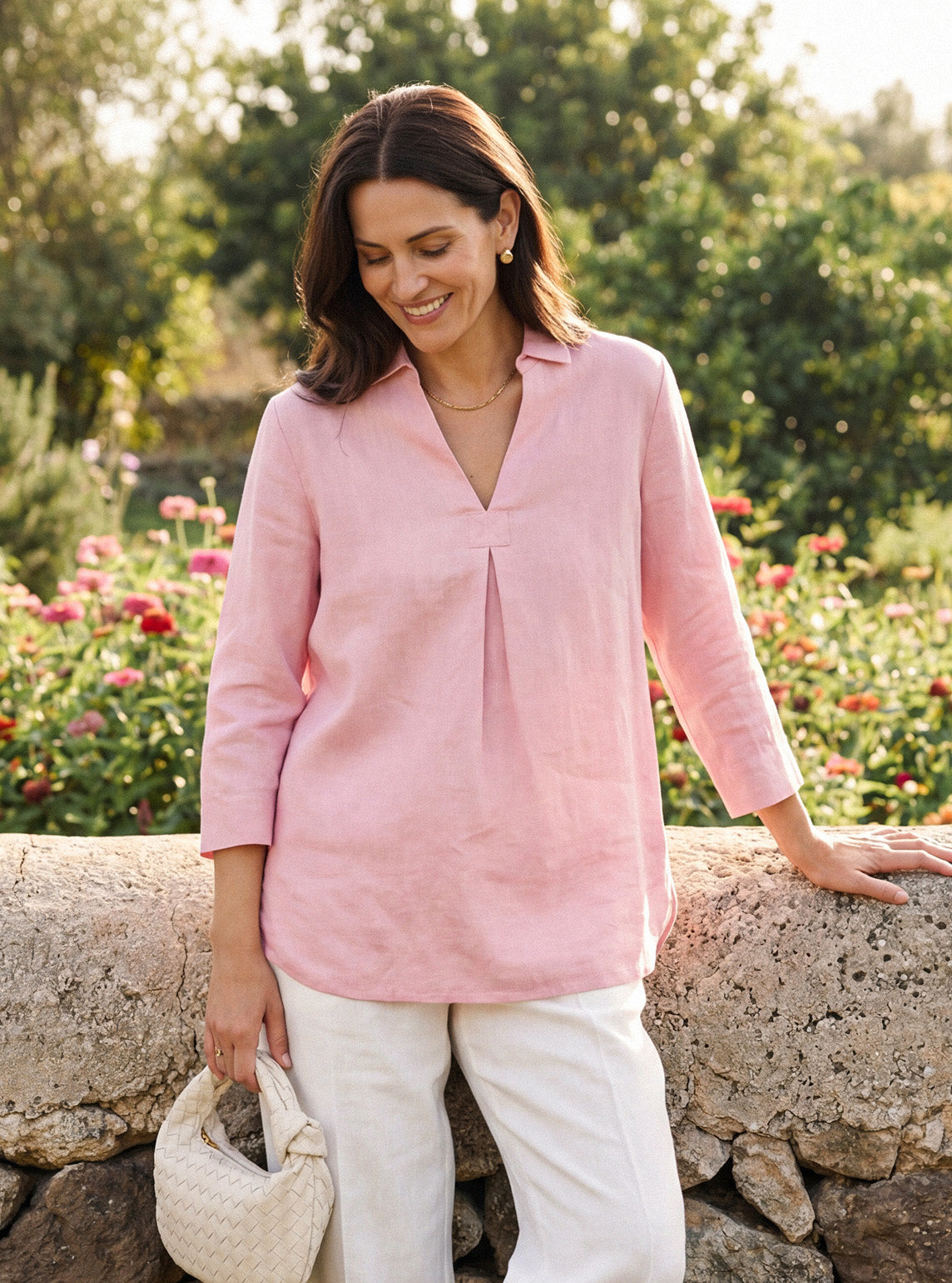 Woman wearing a pink blouse and white pants standing outdoors with flowers and greenery in the background