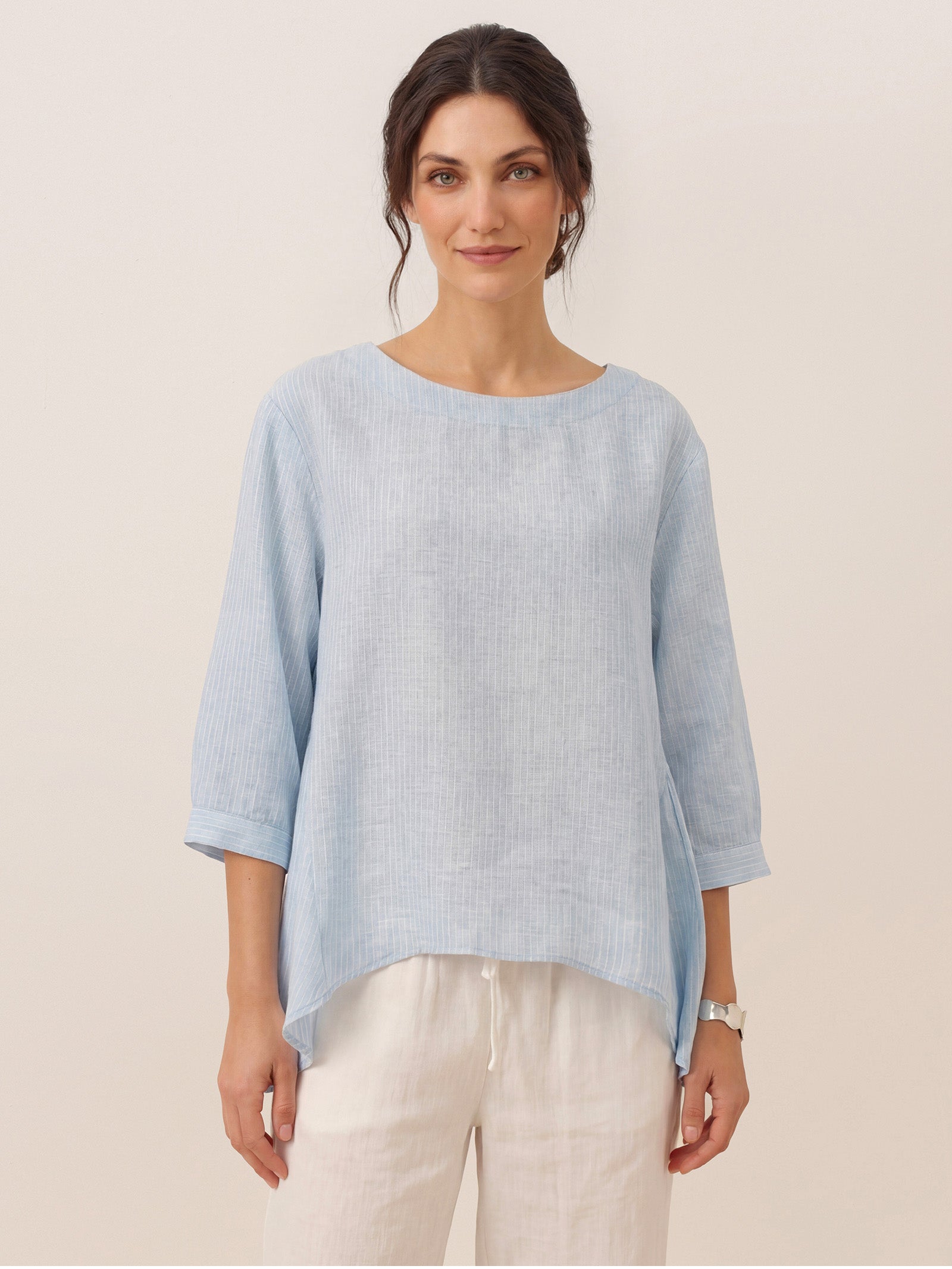 Woman wearing a light blue sweater over a white shirt with beige pants on a plain background