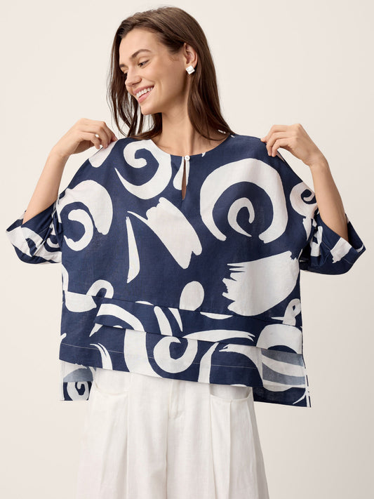 A woman wearing a navy blue and white abstract print top with elbow-length sleeves and a split neckline, paired with white pants, against a solid light beige background.