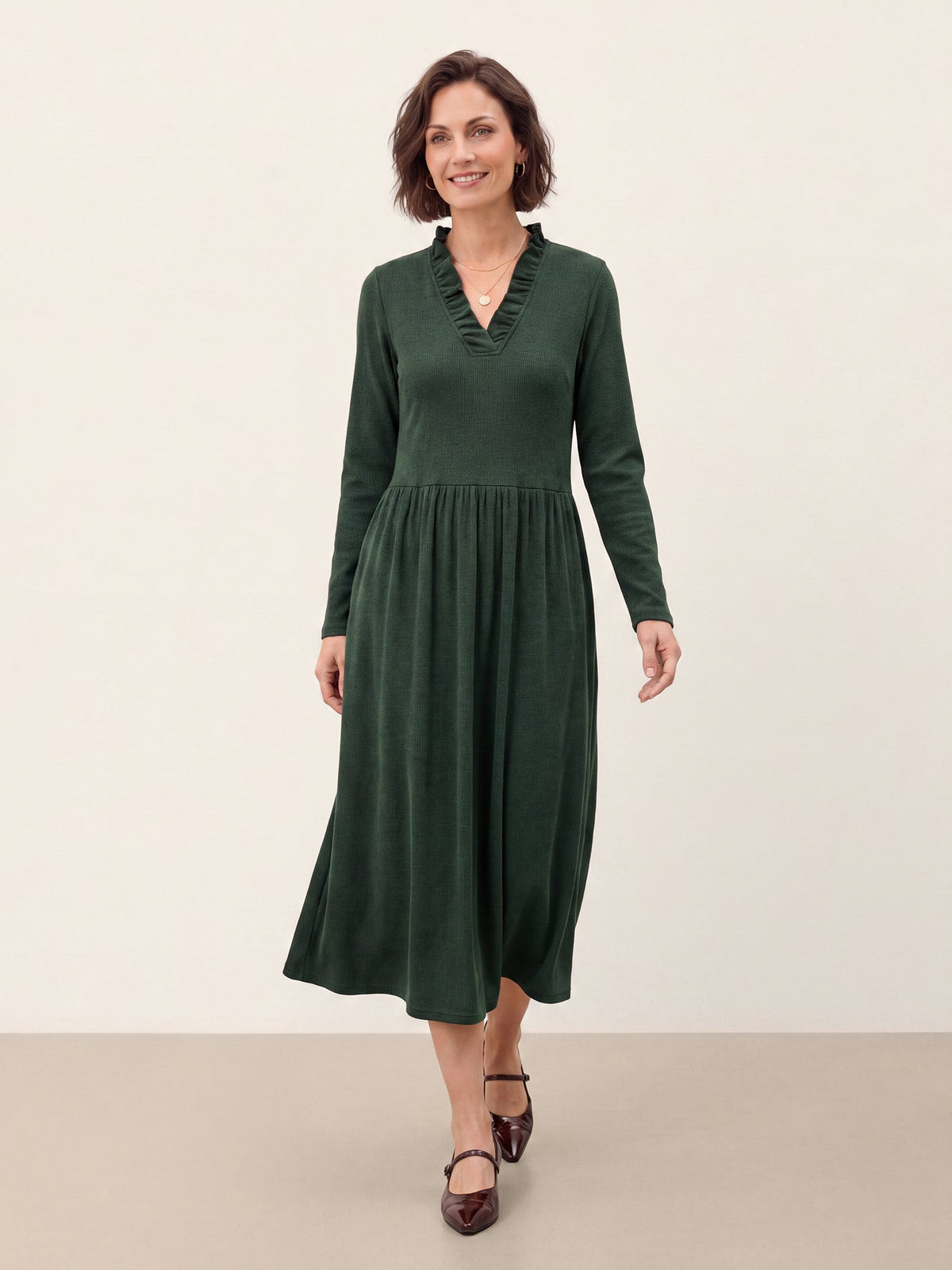 A woman modeling a dark green long-sleeve midi dress with a ruffled v-neckline, standing against a plain beige studio background.