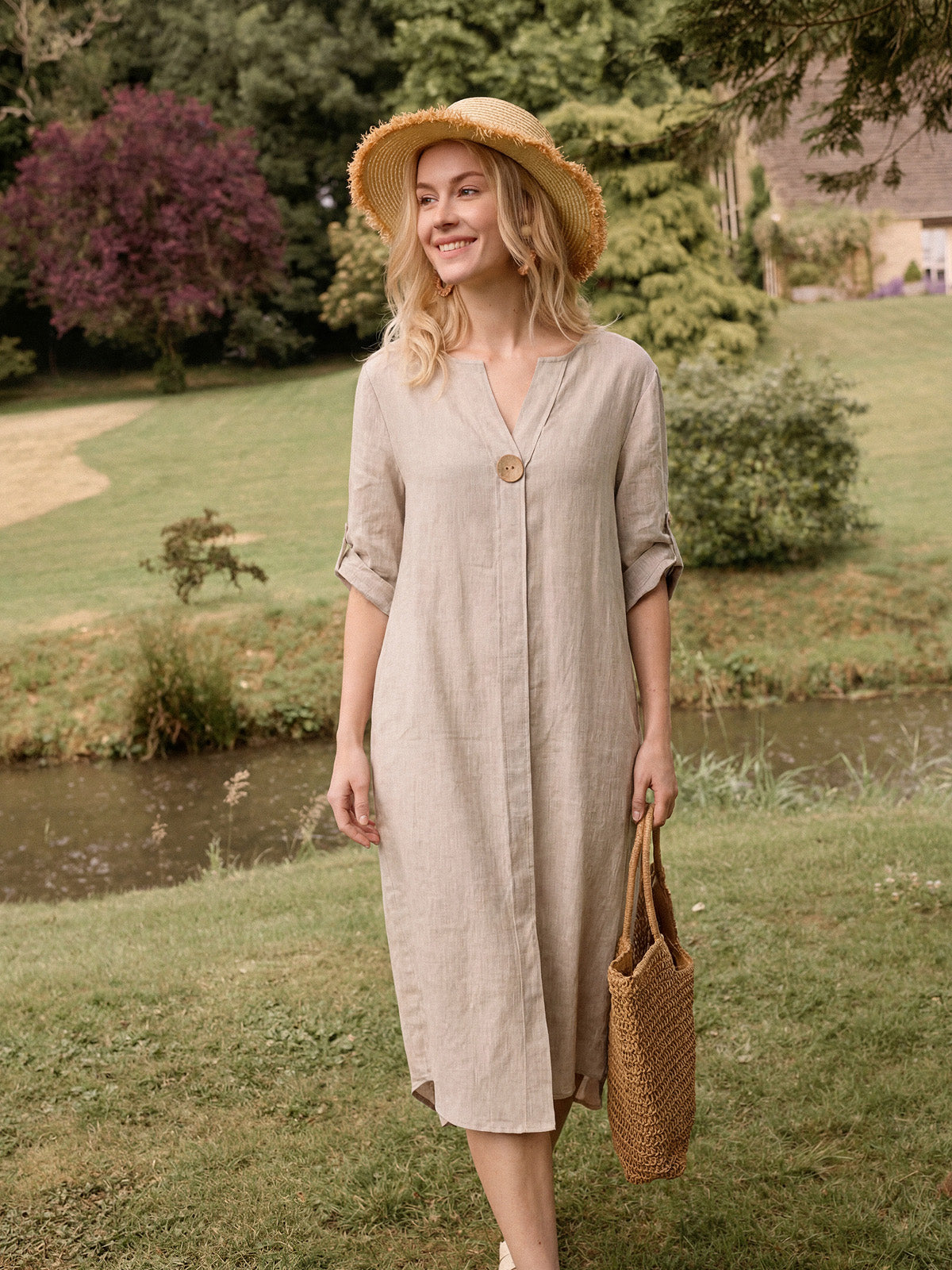 Woman in a beige dress and straw hat standing by a pond in a garden.