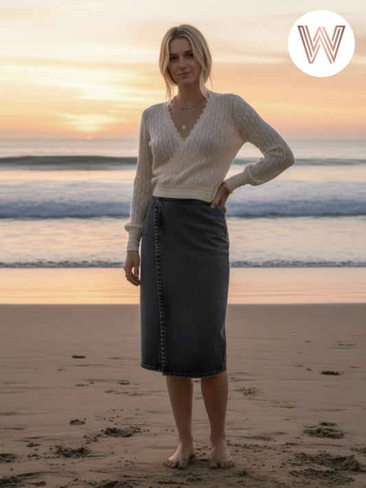 Woman standing on a beach with a sunset in the background, wearing a white sweater and dark skirt.