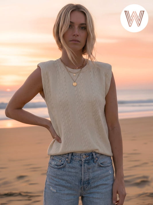 Woman standing on a beach at sunset wearing a beige sleeveless top and blue jeans.