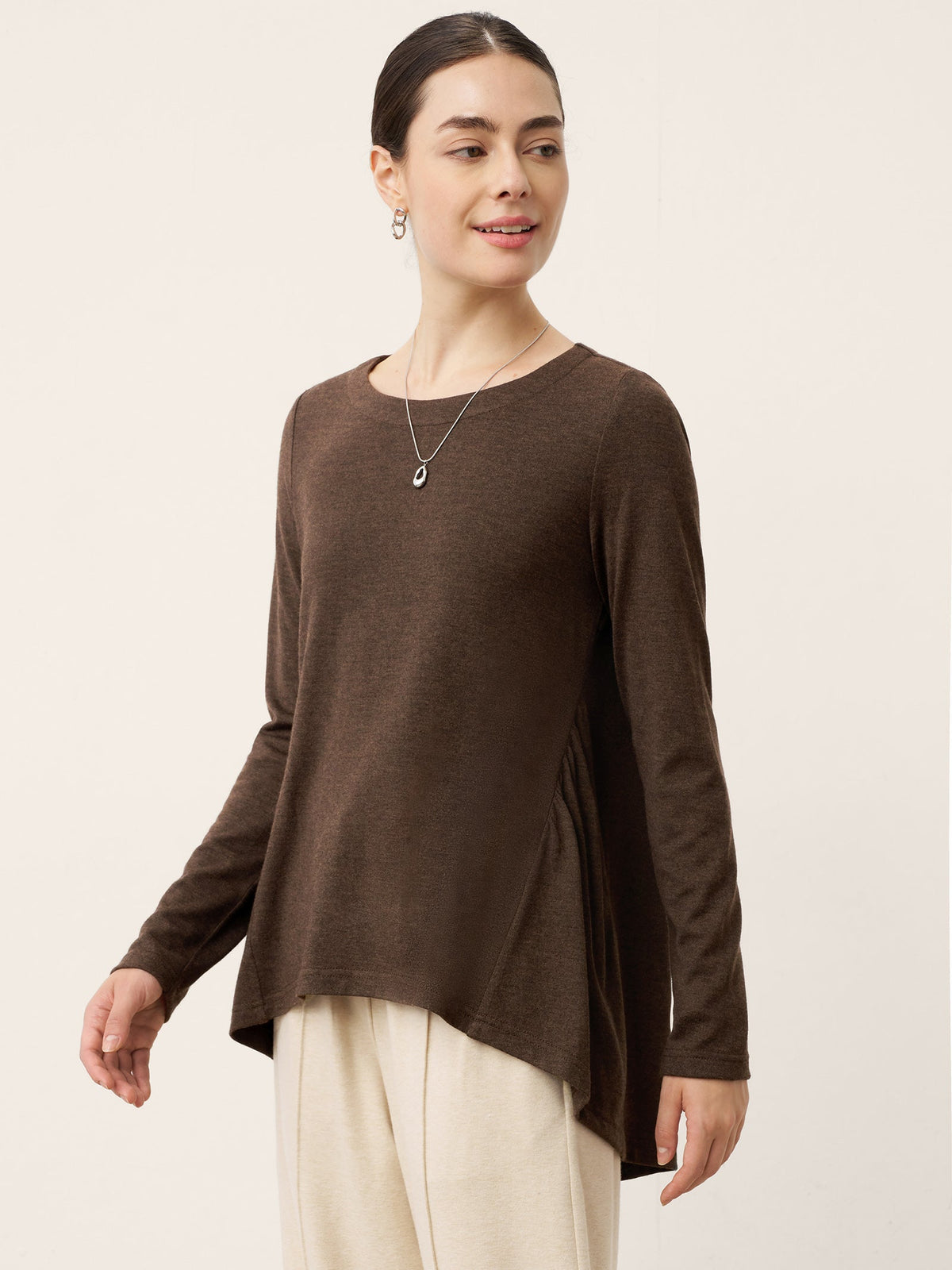 A woman modeling a brown long-sleeve knit top with an asymmetrical draped hem, paired with cream pants, against a plain light beige studio background.