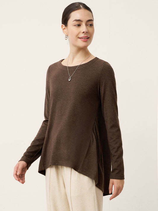 A woman modeling a brown long-sleeve knit top with an asymmetrical draped hem, paired with cream pants, against a plain light beige studio background.