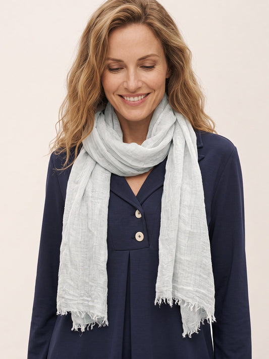 A woman with blonde hair wearing a light gray linen scarf and a navy blue button-up dress, photographed against a solid off-white background.
