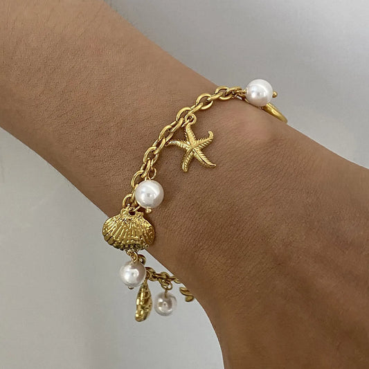 Gold bracelet with starfish, shell, and pearl charms on a wrist against a neutral background