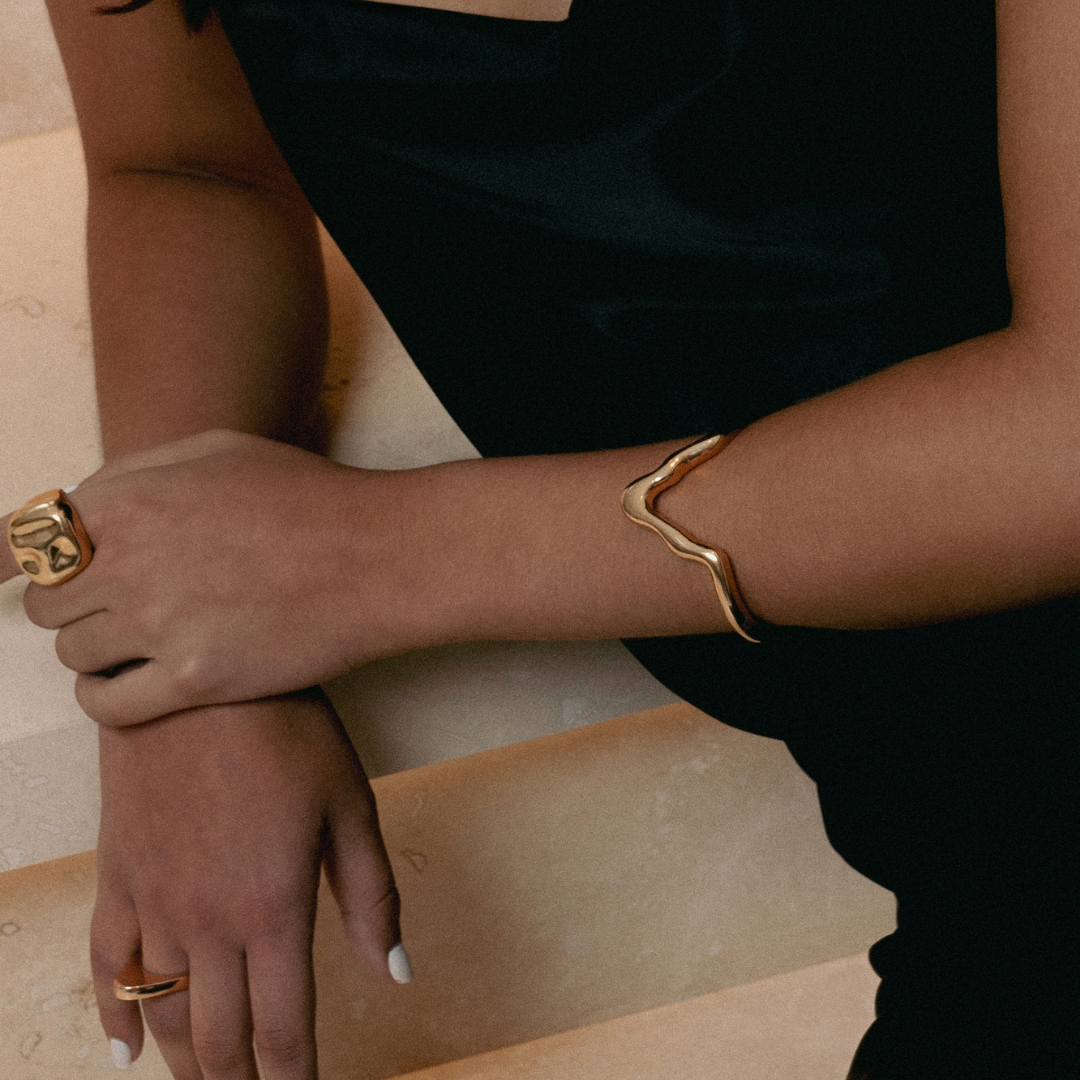 Close-up of a person's arm wearing a gold bracelet and ring on a neutral background