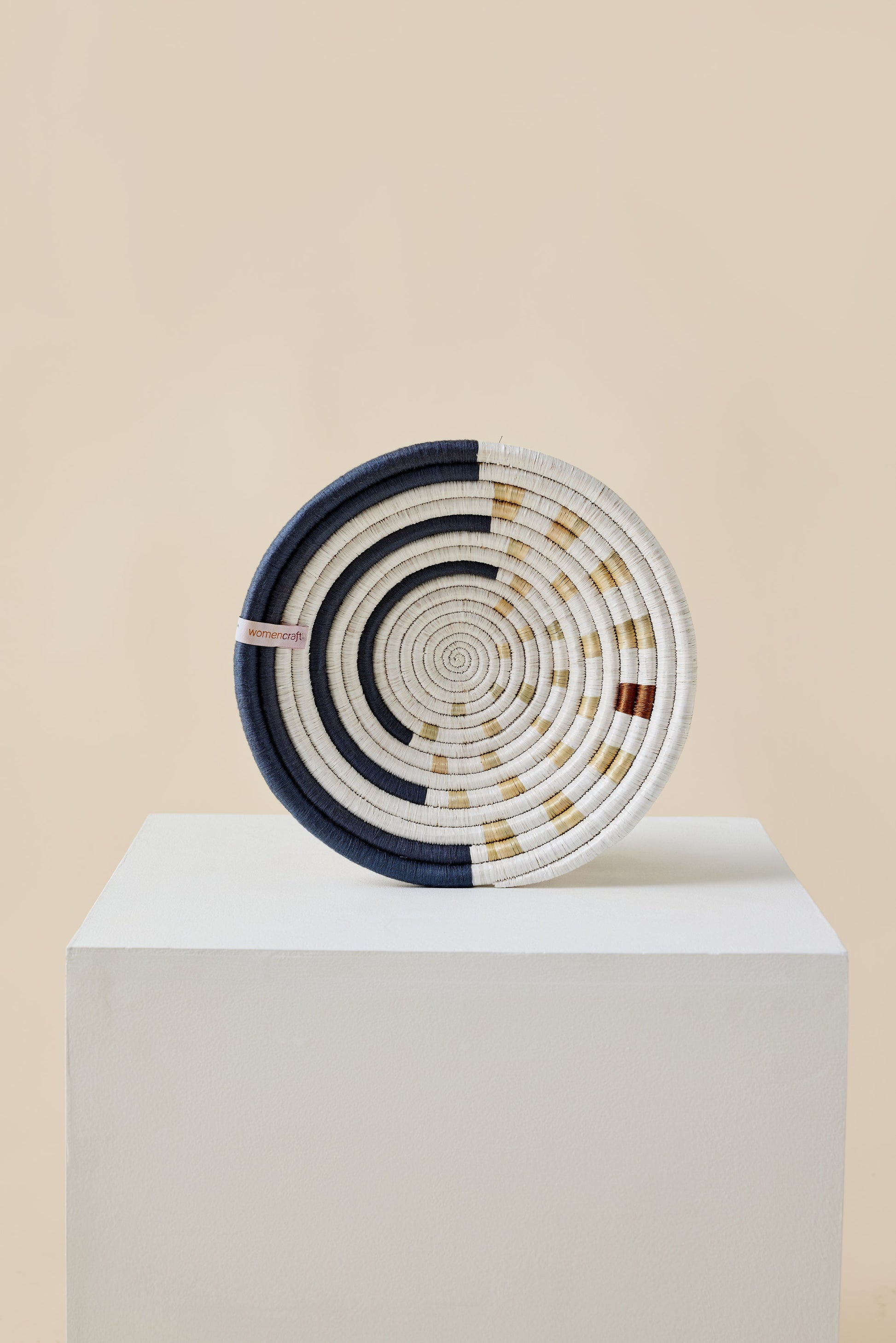 Woven basket with concentric circles on a white pedestal against a beige background