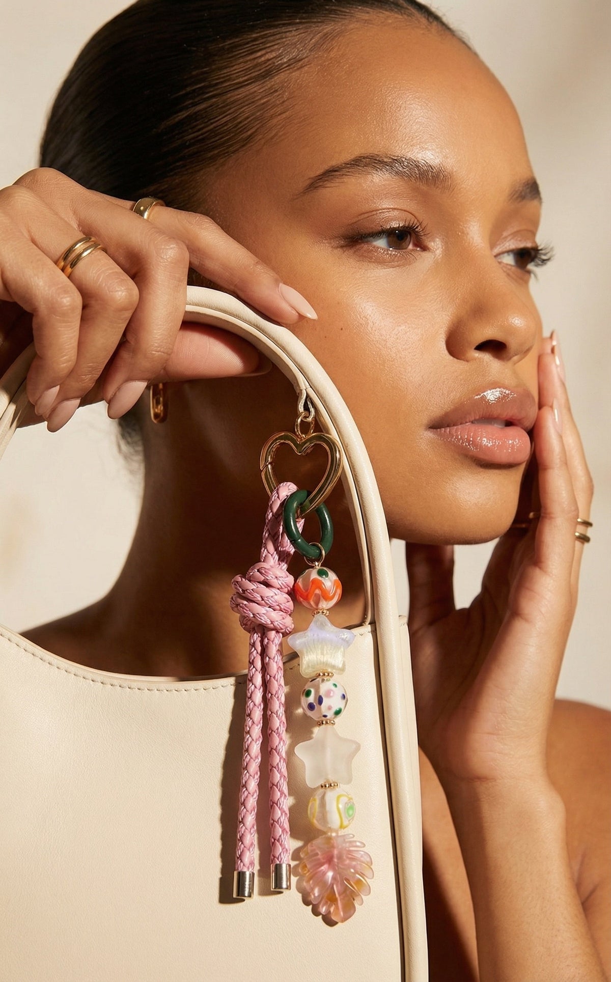 A close-up lifestyle shot of a model wearing a large, whimsical pink and multicolor tassel earring with charms, holding a cream-colored shoulder bag.